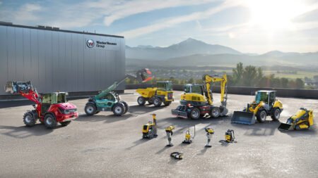 Wacker-Neuson-Group_H1jpg - Machines Wiki Industrial yard with yellow, red, and green construction machines parked beside a building bearing the Wacker Neuson Group sign; mountains in the background.