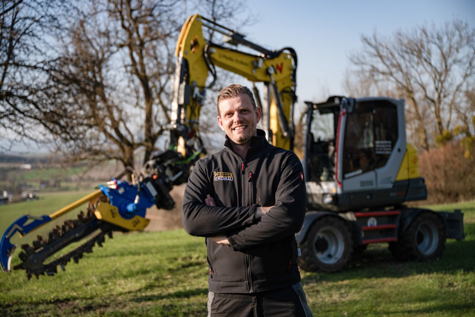Wacker-Neuson_EW100_Pfeffer-Erdbaujpg - Machines Wiki Smiling man in a black jacket with arms crossed, standing in a field with a large yellow excavator behind him.