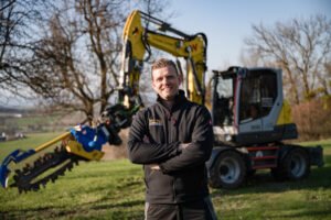 Wacker-Neuson_EW100_Pfeffer-Erdbaujpg - Machines Wiki Smiling man in a black jacket with arms crossed, standing in a field with a large yellow excavator behind him.