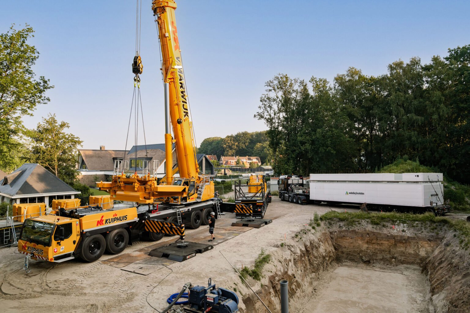 Large yellow construction crane mounted on a multi-axle truck at a residential construction site, with a deep excavation pit in the foreground and houses and trees in the background.