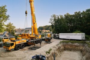 Large yellow construction crane mounted on a multi-axle truck at a residential construction site, with a deep excavation pit in the foreground and houses and trees in the background.