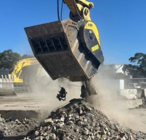 Yellow excavator bucket breaking concrete rubble at a construction site, dust rising as debris falls from the bucket.